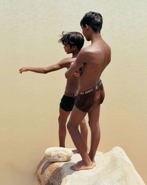 Two bare-chested boys stand on a rock in a river, one with arms outstretched, the other looking on, captured in a medium outdoor shot.