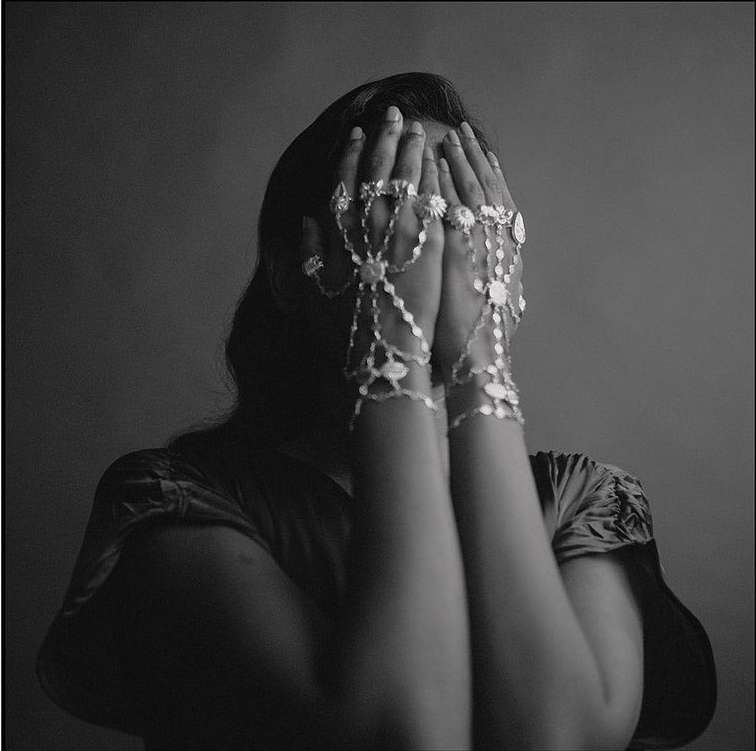 Adult female in a dark gray studio hiding her face with both hands. She wears a silver hand flower bracelet.