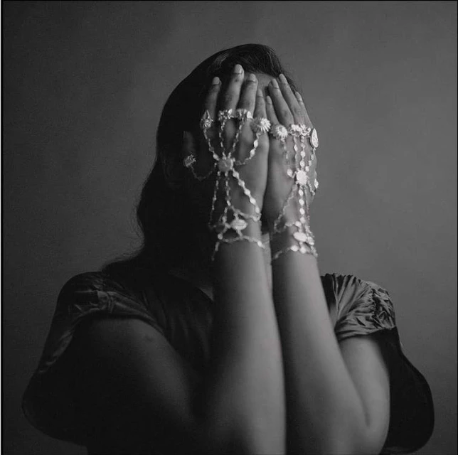 Adult female in a dark gray studio hiding her face with both hands. She wears a silver hand flower bracelet.