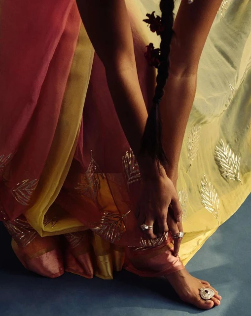 Woman's foot and hands adorned with jewelry and a floral braid, set against a blue backdrop with a flowing pastel saree.