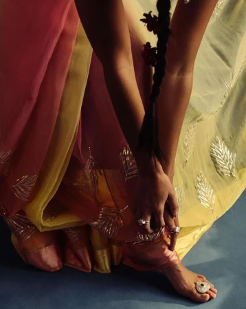 Woman's foot and hands adorned with jewelry and a floral braid, set against a blue backdrop with a flowing pastel saree.