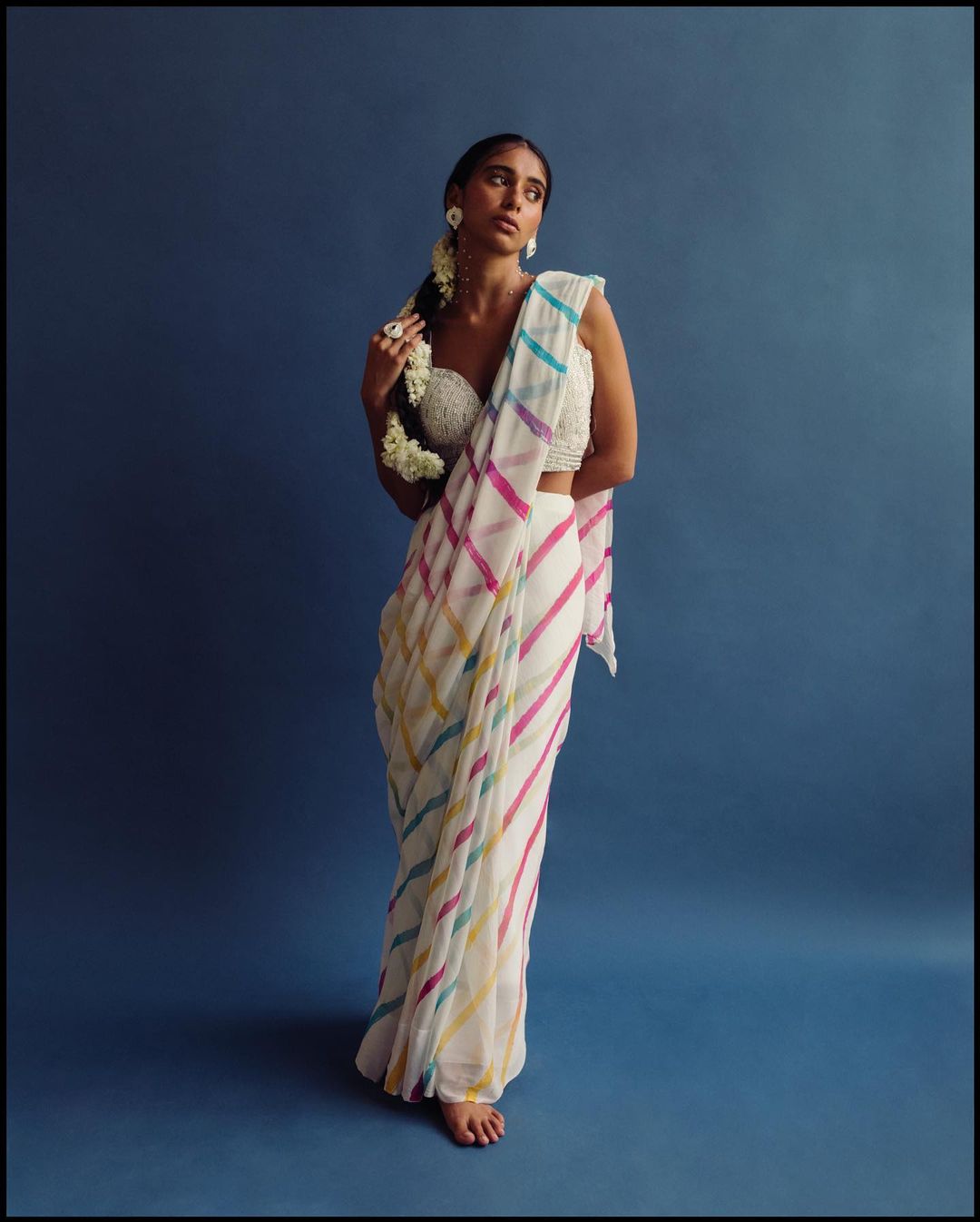 An adult female model poses in a white sari with colorful stripes, adorned with flowers, against a dark blue studio backdrop.