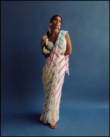 An adult female model poses in a white sari with colorful stripes, adorned with flowers, against a dark blue studio backdrop.