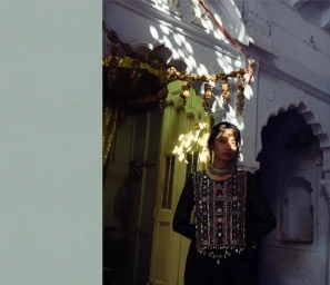 South Asian woman in embroidered black tunic standing in doorway with dappled sunlight by Tenzin Lhagyal