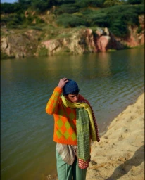 Man wearing a vibrant orange patterned sweater and blue beanie standing by a calm lake with sandy banks.