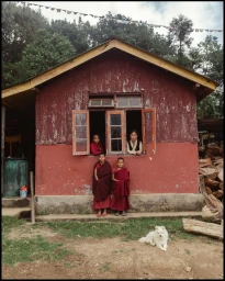 Four children and a woman posing outside a red wooden house with a white dog resting in the foreground.