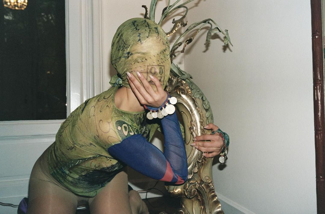 A person in a patterned green shirt and face covering kneels, resting their hand on their face and leaning against a decorative gold chair.