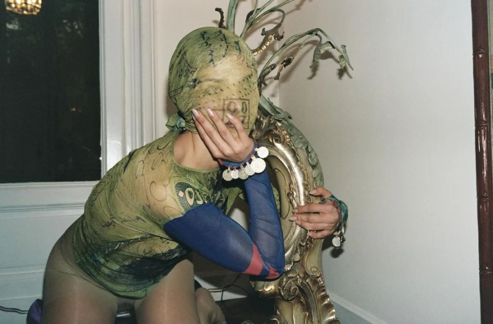 A person in a patterned green shirt and face covering kneels, resting their hand on their face and leaning against a decorative gold chair.