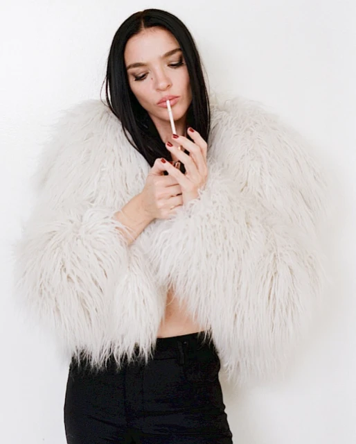 Elegant woman in a furry white coat holds a cigarette in an indoor shot. She wears black pants with a blank background.