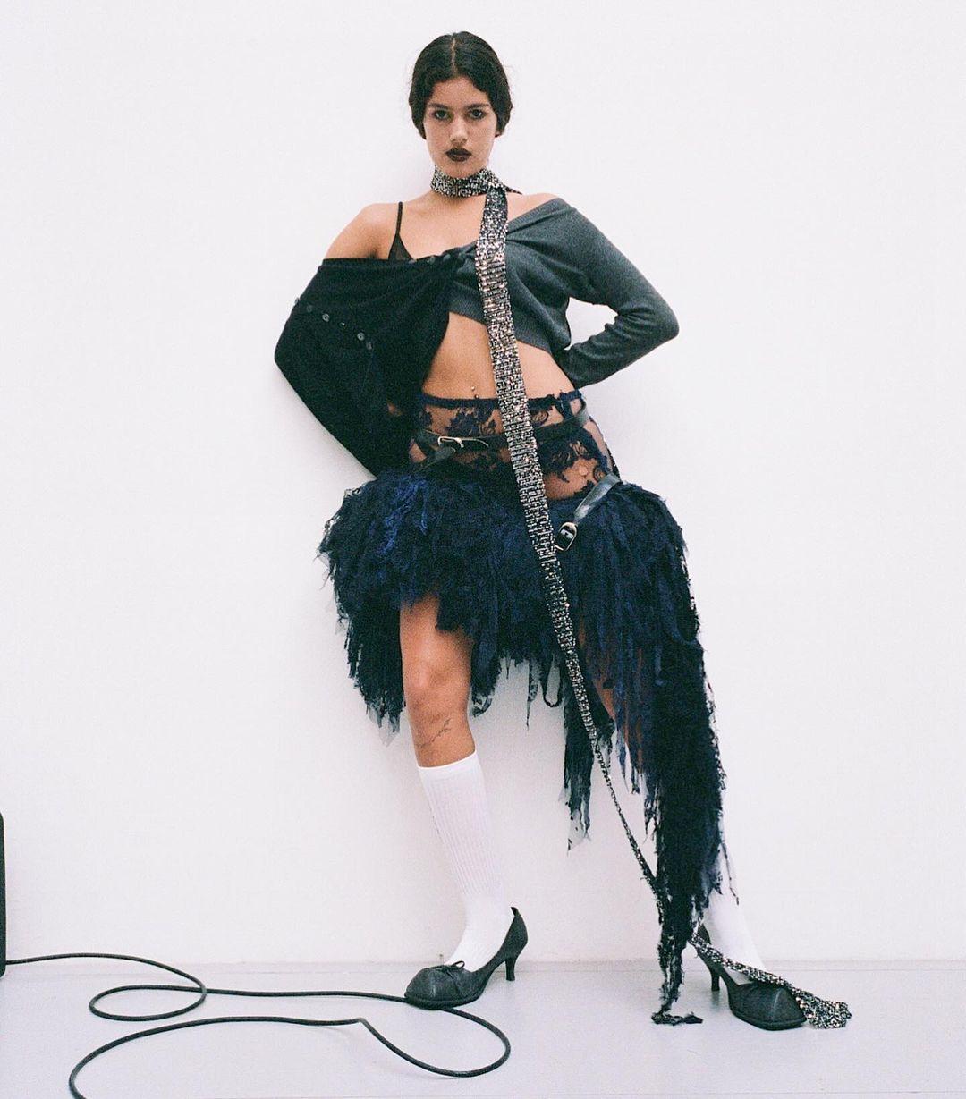 A young woman stands in a fashion pose in a studio. She wears a knit top, a lace skirt with fringe, white socks, and heels.