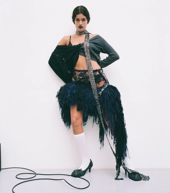 A young woman stands in a fashion pose in a studio. She wears a knit top, a lace skirt with fringe, white socks, and heels.