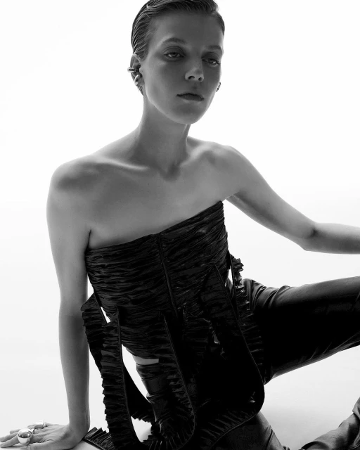 Fashionable woman with slicked-back hair poses in a black, strapless, ruched top with a ruffled skirt and leather pants against a white background.