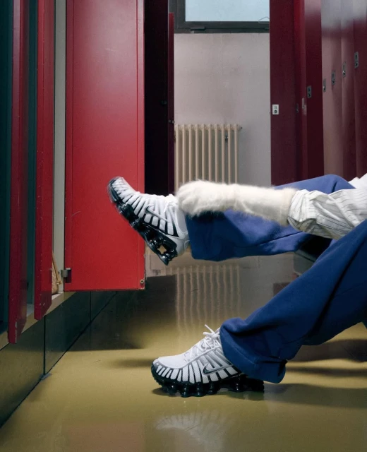 Indoor image featuring a person reclining with legs up against red lockers wearing white sneakers with black soles in a locker room.