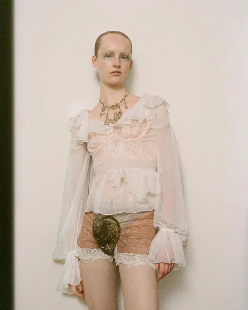 Adult female model in sheer ruffled blouse and shorts with shell detail, standing against a neutral wall in a fashion editorial.