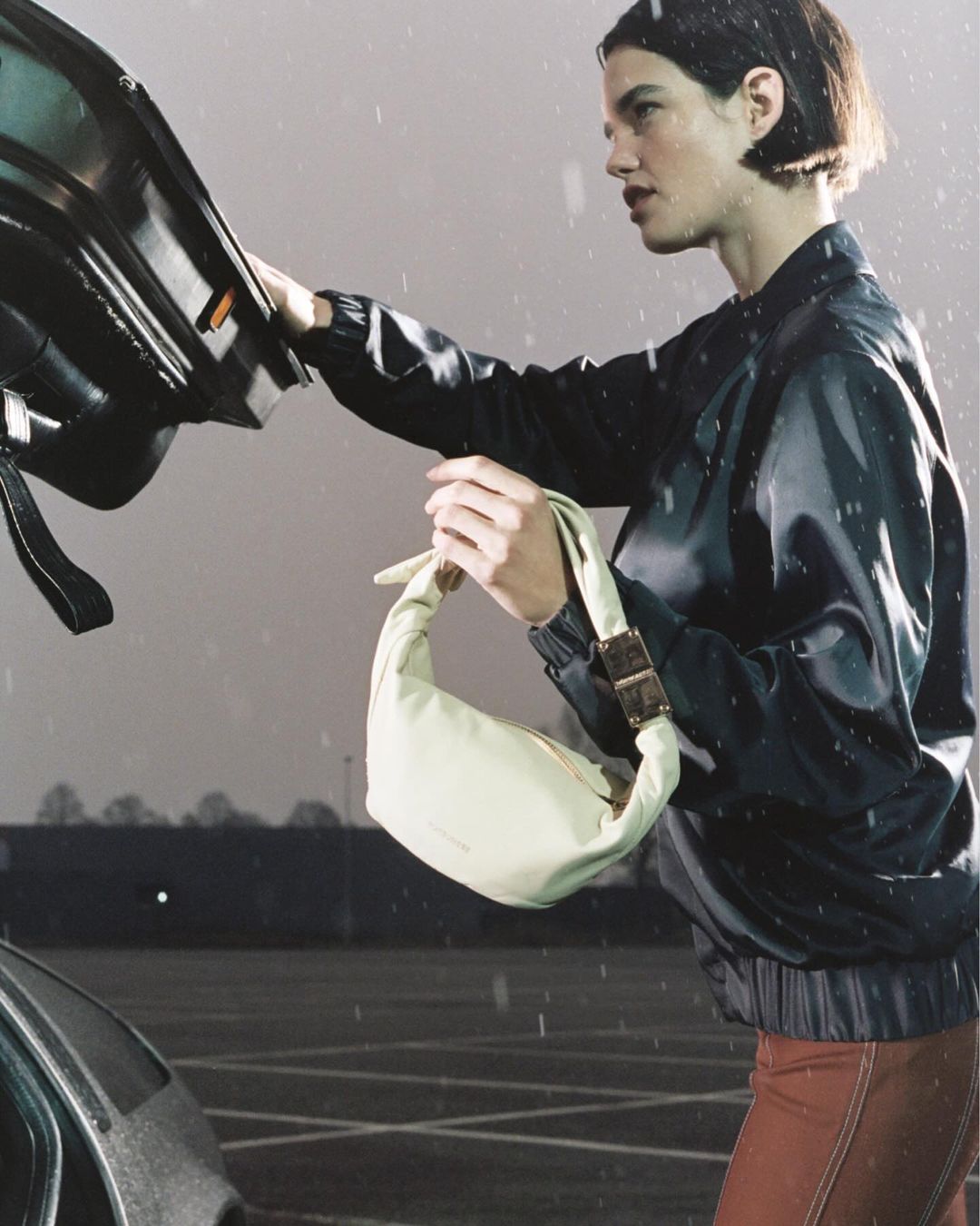 Adult female carrying a light-yellow handbag next to a black car, in a wet parking lot with a gray sky.