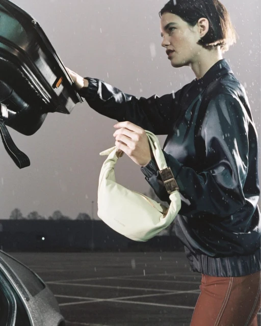 Adult female carrying a light-yellow handbag next to a black car, in a wet parking lot with a gray sky.