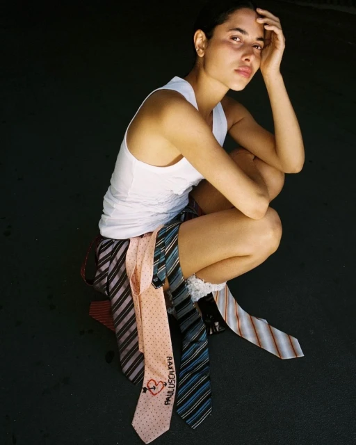 Woman in white top and striped skirt poses in a crouch with multiple neckties draped around her legs in a softly lit studio setting.