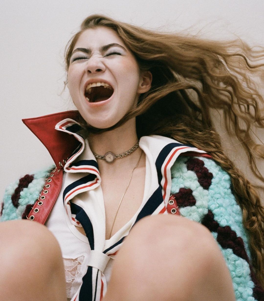 Close-up of a young woman with long hair, wearing a nautical-themed jacket and jewelry, with her mouth open in an expressive yell.