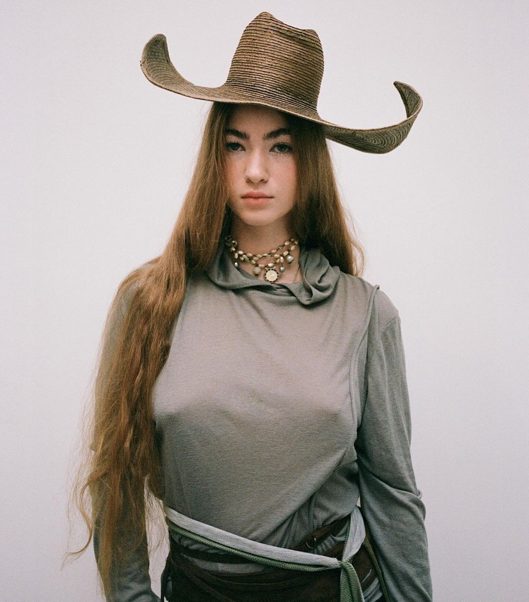 Portrait of a young woman with long brown hair wearing a wide-brimmed woven hat, layered sheer top, and beaded necklace against a white background.