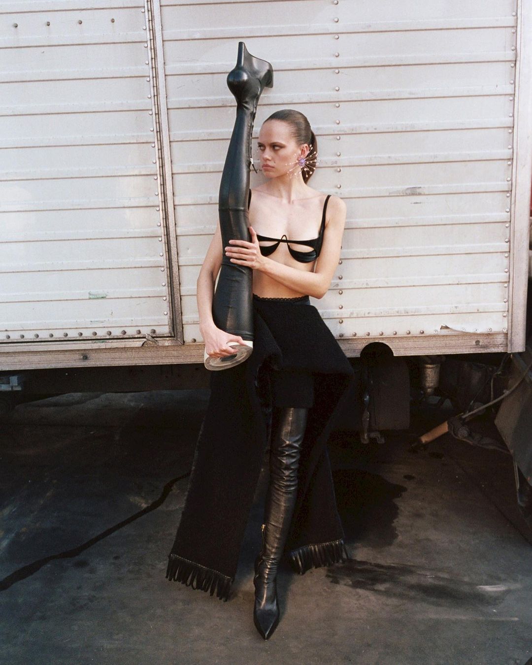 Fashion model posing with an artificial leg and thigh-high leather boot against a corrugated metal background in a studio setting.