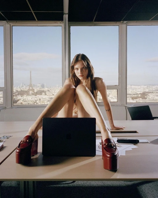 Fashion editorial shot of a young adult woman posing on a desk with platform shoes and a laptop against a city skyline backdrop.