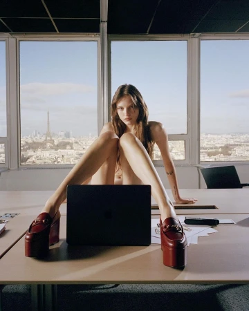 Fashion editorial shot of a young adult woman posing on a desk with platform shoes and a laptop against a city skyline backdrop.