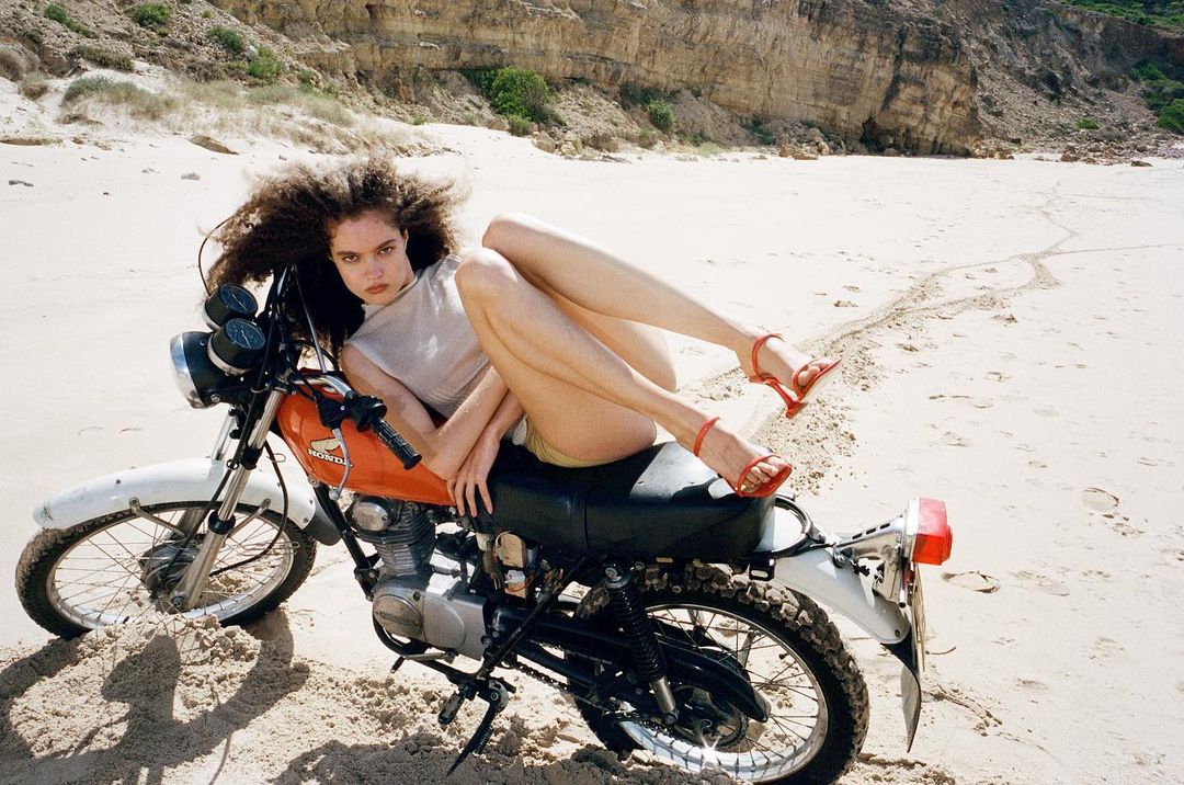 Woman with curly hair poses on a vintage motorcycle on a sandy beach with cliffs in the background in an outdoor fashion shot.