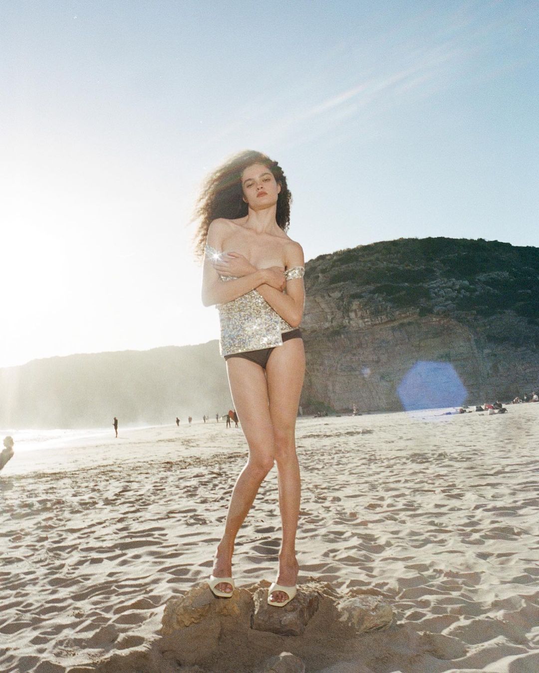 Model on a sunny beach with a shimmering top and dark bikini bottom, accessorized with jewelry and heels, posing on a sandy shore.