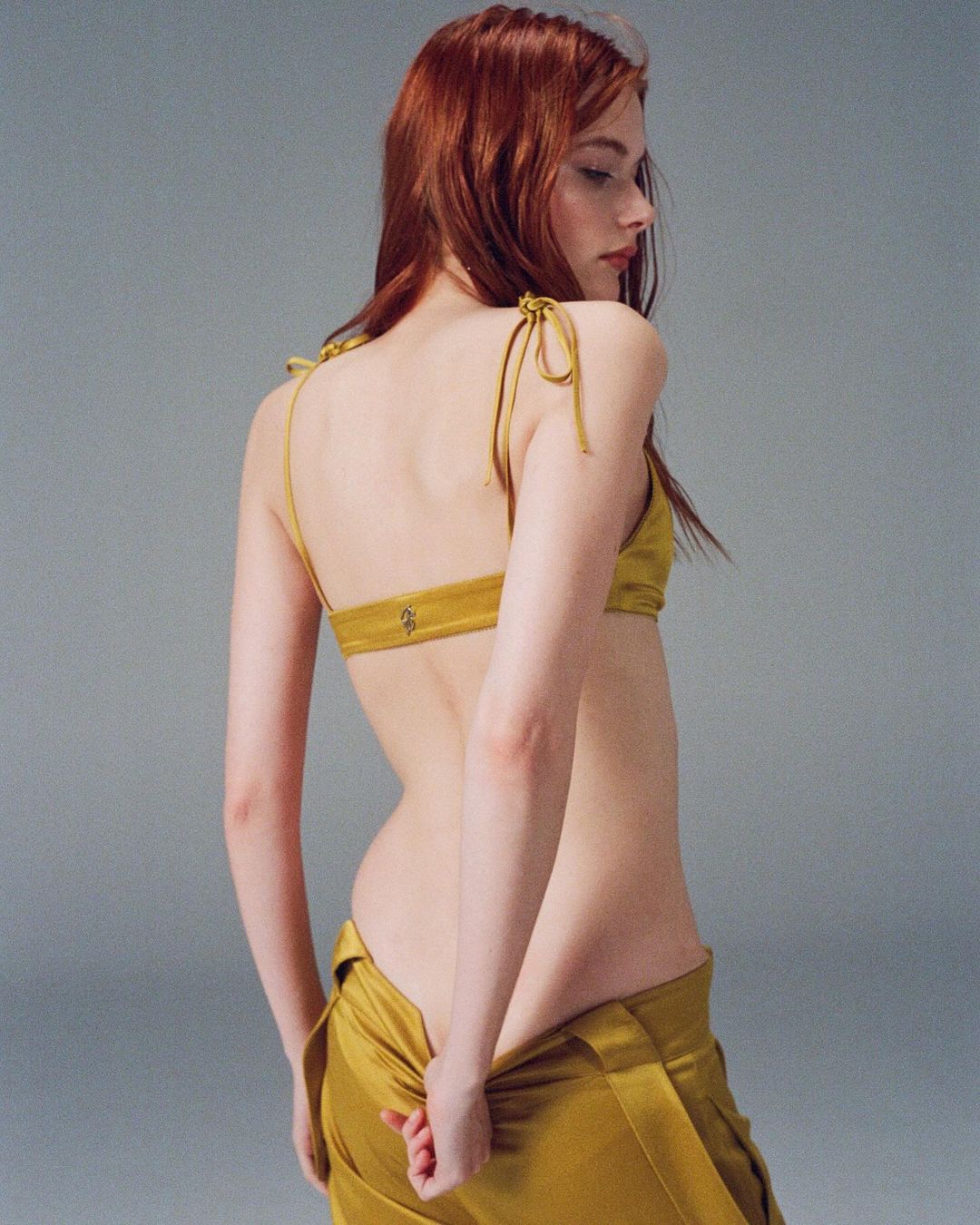 A fair-skinned woman with auburn hair poses in a mustard-yellow top and pants, showcasing a fashion-forward look against a neutral backdrop.