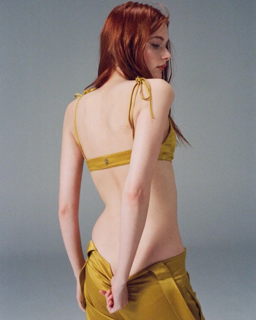 A fair-skinned woman with auburn hair poses in a mustard-yellow top and pants, showcasing a fashion-forward look against a neutral backdrop.