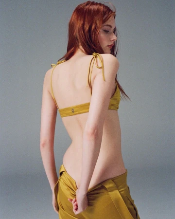 A fair-skinned woman with auburn hair poses in a mustard-yellow top and pants, showcasing a fashion-forward look against a neutral backdrop.