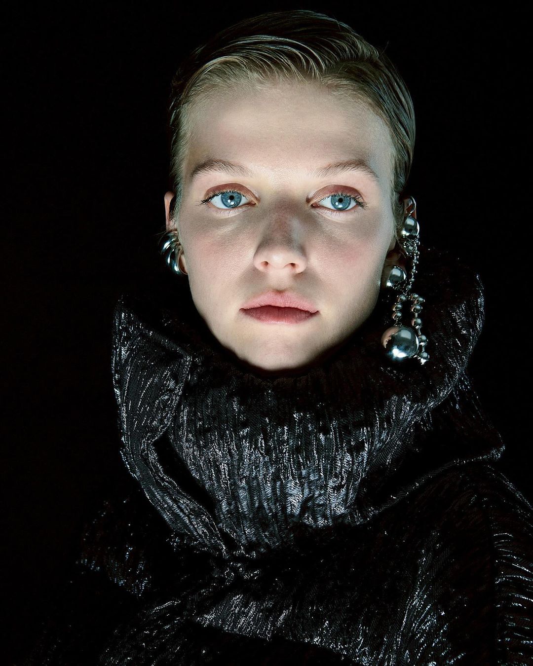 Close-up portrait of a woman with slicked hair wearing metallic earrings and a black textured high-collar coat against a dark background