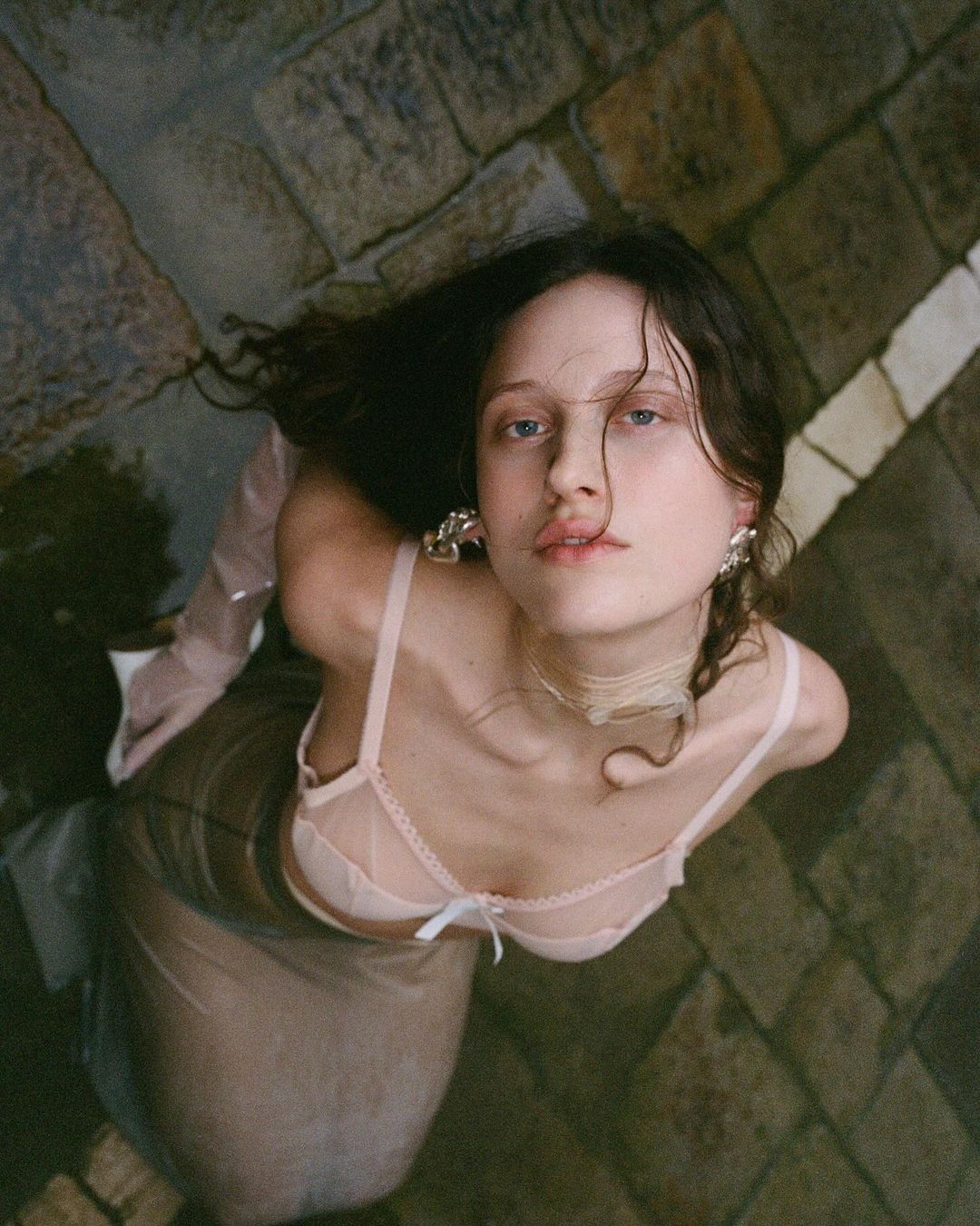 High angle shot of a woman with dark hair wearing a pale pink bra and sheer skirt, looking up while positioned on stone paving.