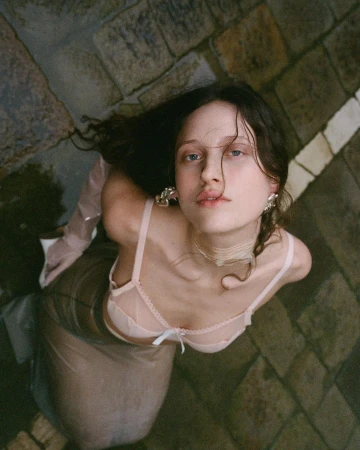 High angle shot of a woman with dark hair wearing a pale pink bra and sheer skirt, looking up while positioned on stone paving.
