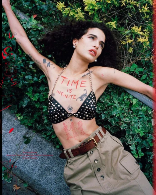 Woman lying on a stone structure near green foliage, wearing a studded top and high-waisted khaki pants with a brown belt and tattoos.