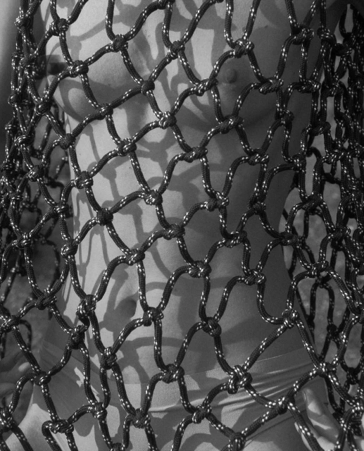 Black and white close-up of an adult with a net shirt, casting shadows. The image is moody and textural, and focuses on the details.