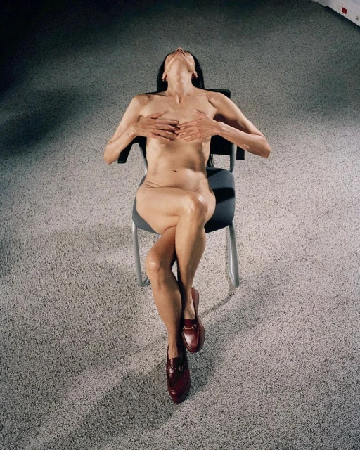A woman sits on a chair with legs crossed, chest covered by her hands, and head tilted back in an indoor setting with grey carpet.