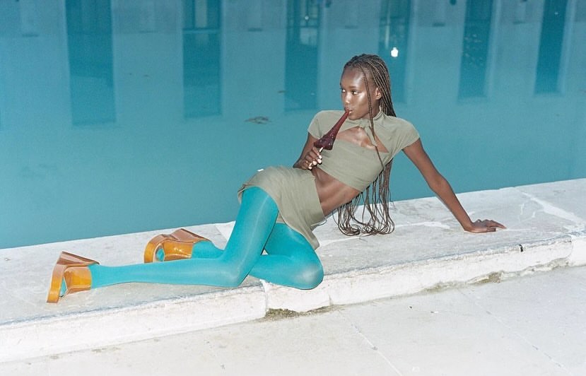 Woman poses in sage green crop top and mini skirt with blue tights next to a pool, holding a dark brown glass bottle.