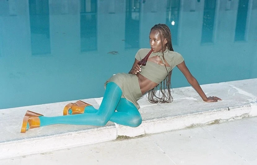 Woman poses in sage green crop top and mini skirt with blue tights next to a pool, holding a dark brown glass bottle.