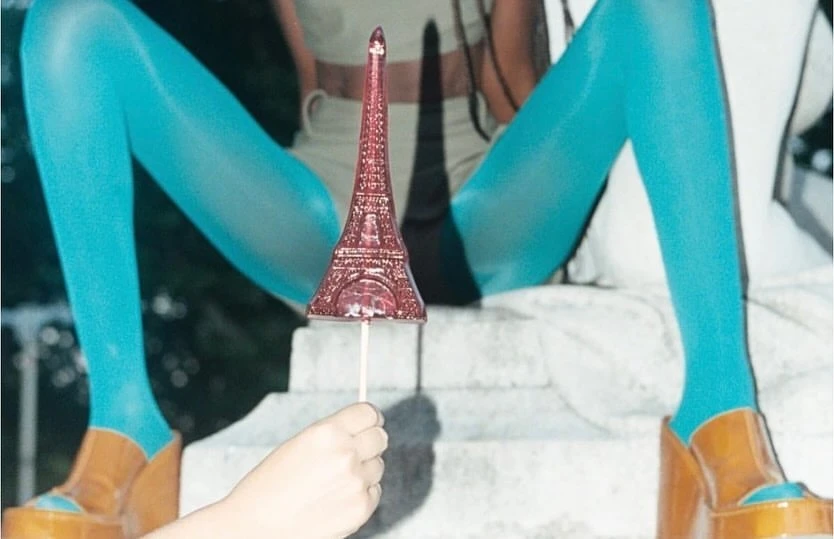 Person in aqua tights sitting with brown platform shoes, holding a small Eiffel Tower souvenir against an out-of-focus background.