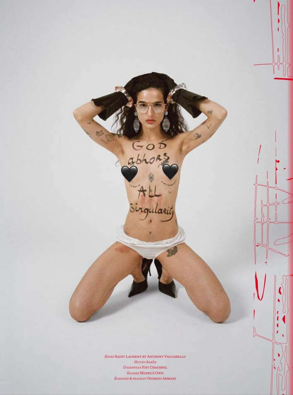 Woman kneels in a photo studio with handwritten text on her body, glasses, jewelry, black heeled shoes, and abstract red markings.