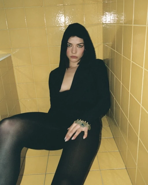 Woman in black hooded outfit seated in tiled bathroom with hand jewelry looking at camera; yellow tiled walls and flooring; indoor lighting.