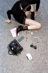Woman in black sitting on pavement surrounded by dropped accessories including a handbag, smartphone, sunglasses, and cosmetics.