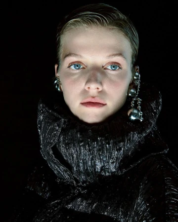 Close-up portrait of a woman with slicked hair wearing metallic earrings and a black textured high-collar coat against a dark background