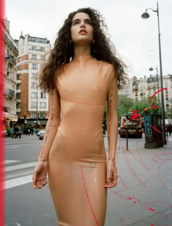 Woman with long dark curly hair wearing a translucent nude-coloured dress standing outdoors on a city street.