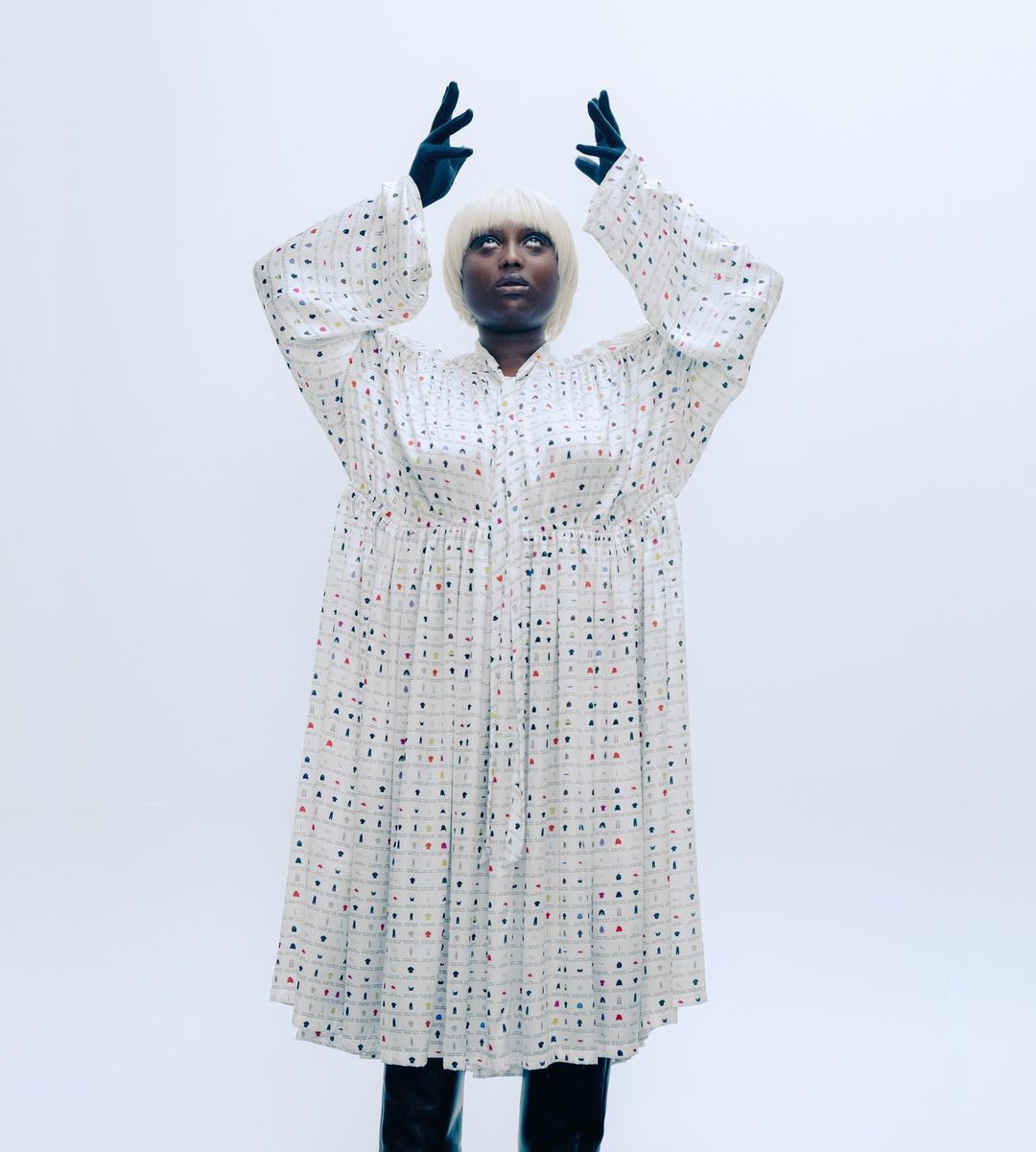 An adult female wearing a long patterned dress, black gloves, and boots stands in a photo studio with arms raised above her head.