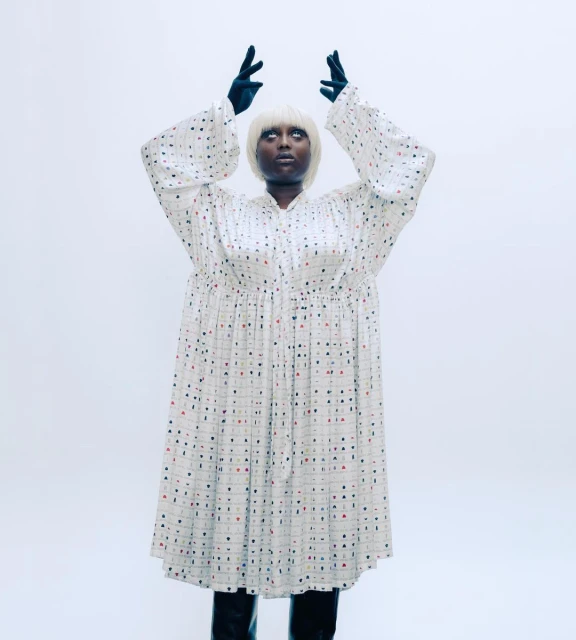 An adult female wearing a long patterned dress, black gloves, and boots stands in a photo studio with arms raised above her head.