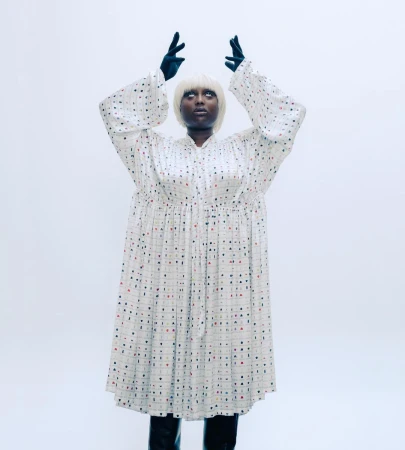 An adult female wearing a long patterned dress, black gloves, and boots stands in a photo studio with arms raised above her head.