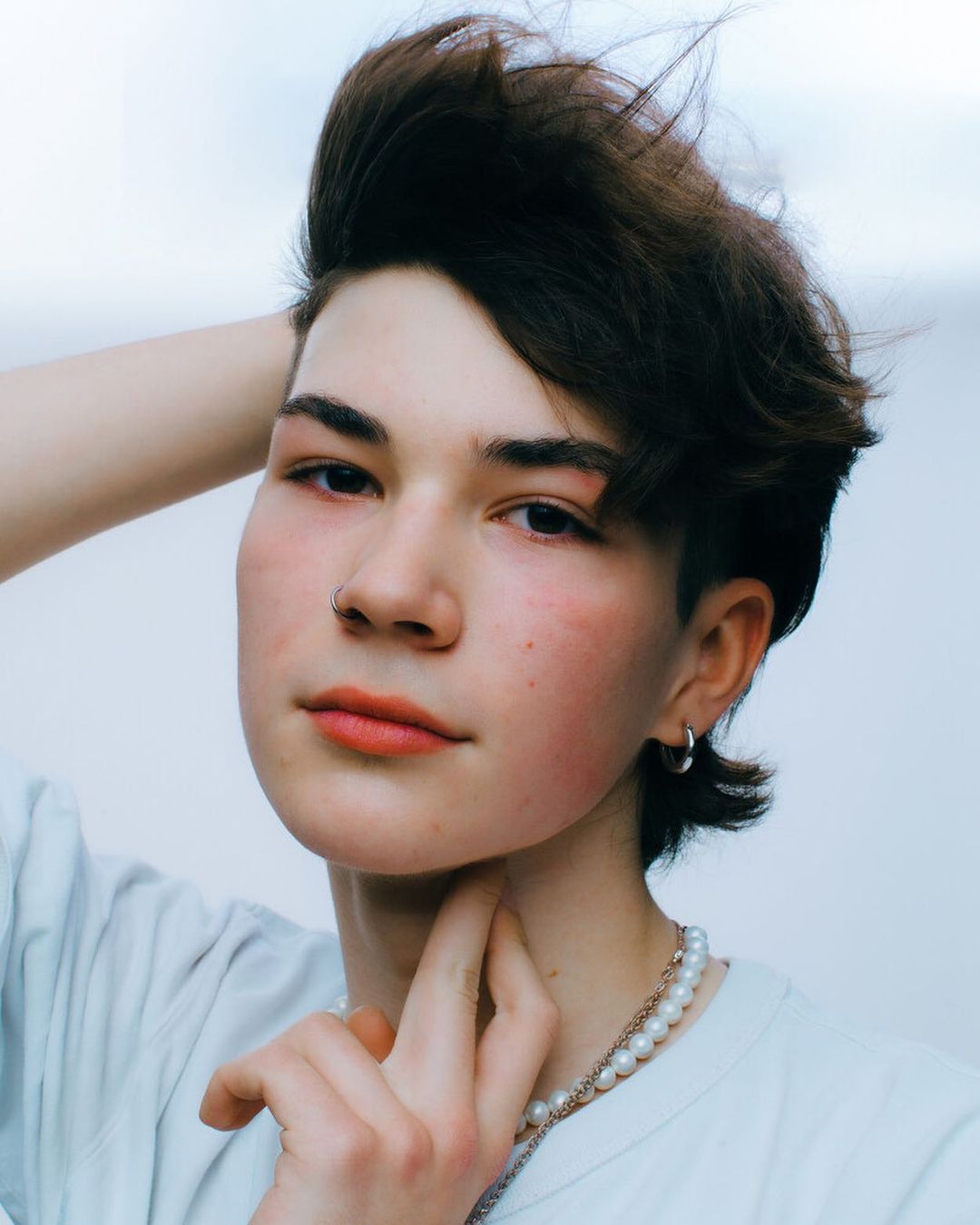 Close-up portrait of a young person wearing a pearl necklace, nose ring, and hoop earring against a plain background in Paris