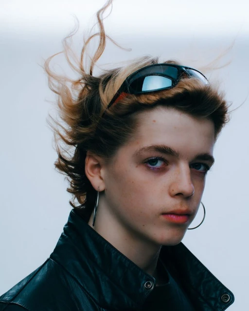 Close-up studio portrait of a young adult with sunglasses on their head, hoop earrings, and a black leather jacket against a light backdrop.